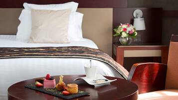 A hotel room with a neatly made bed, a small table with a dessert tray, a white cup, and a vase with flowers on a nightstand.