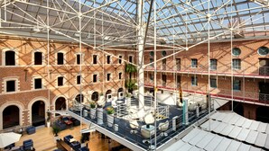 A spacious atrium with a high ceiling and a glass roof, featuring a central open area with seating and a stage, surrounded by brick walls and multiple levels.