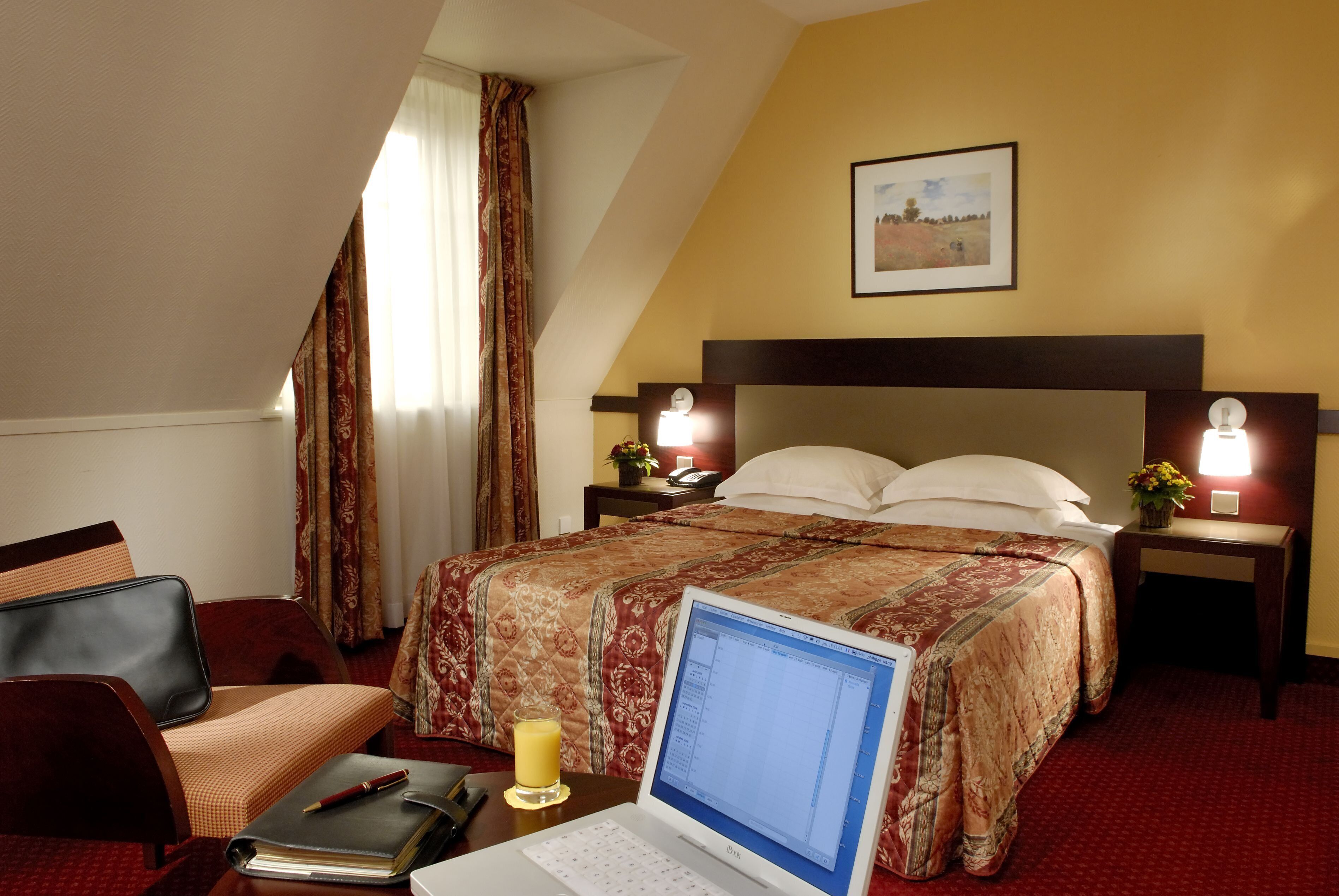 A hotel room with a bed, a desk with a laptop, a chair, a bedside table with a lamp, and a window with curtains.