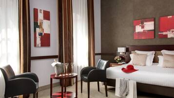 Ein Hotelzimmer mit einem Bett, zwei Stühlen, einem kleinen Tisch mit einer Vase und Gläsern sowie Kunstwerken an den Wänden.