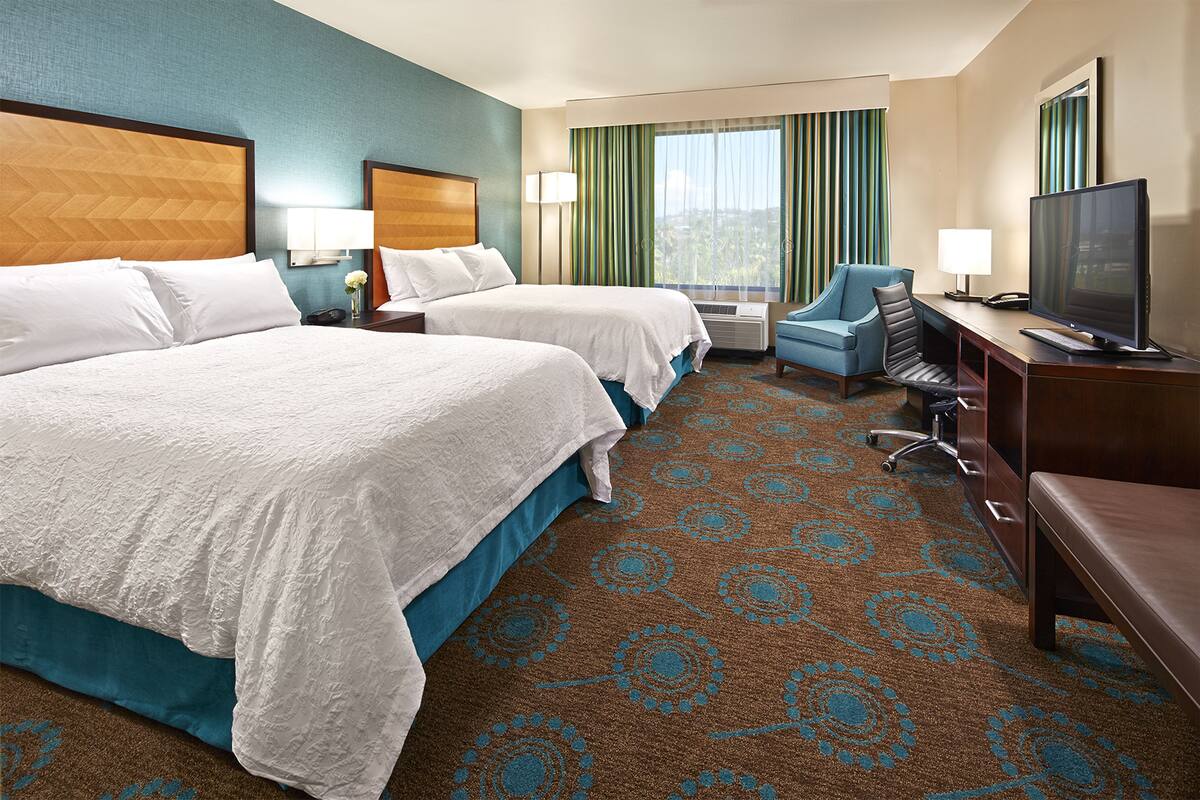 A hotel room with two beds, a desk with a TV, a chair, and a large window with curtains.