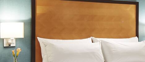 Quarto de hotel com uma cama grande, almofadas brancas, mesa de cabeceira com abajur e um vaso com flores.