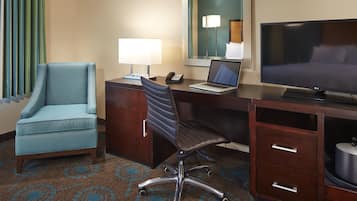 A hotel room with a desk, a laptop, a television, a blue armchair, and a telephone.