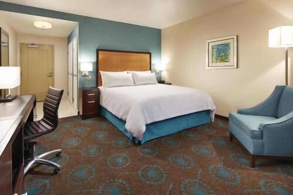 A hotel room with a bed, a desk, a chair, a nightstand, and a wall-mounted lamp.