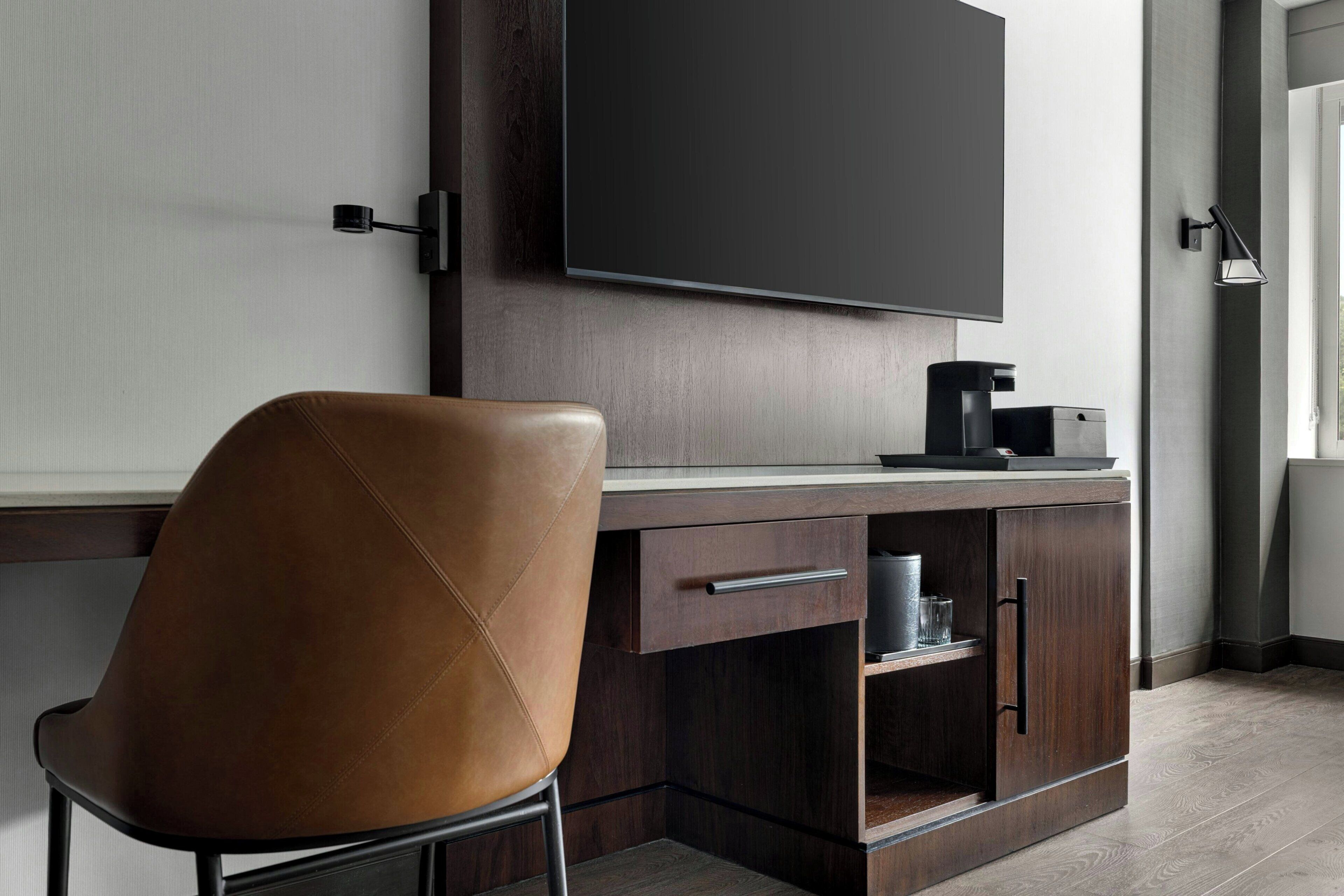 A modern hotel room with a flat-screen TV mounted on the wall, a leather chair, a wooden desk with a coffee maker, and a built-in cabinet.