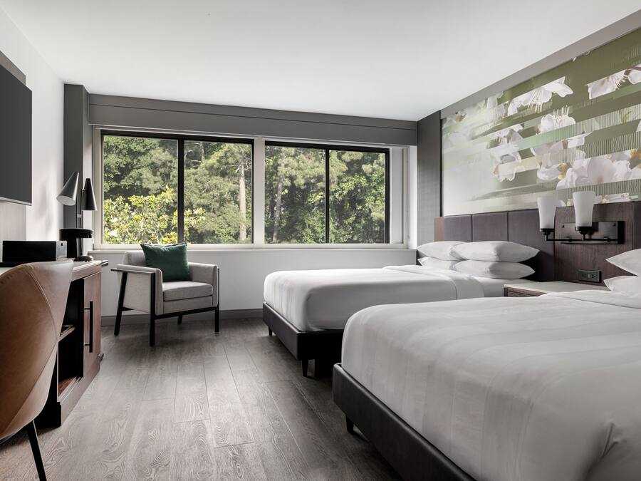 A hotel room with two beds, a desk, a chair, and a large window with a view of trees.
