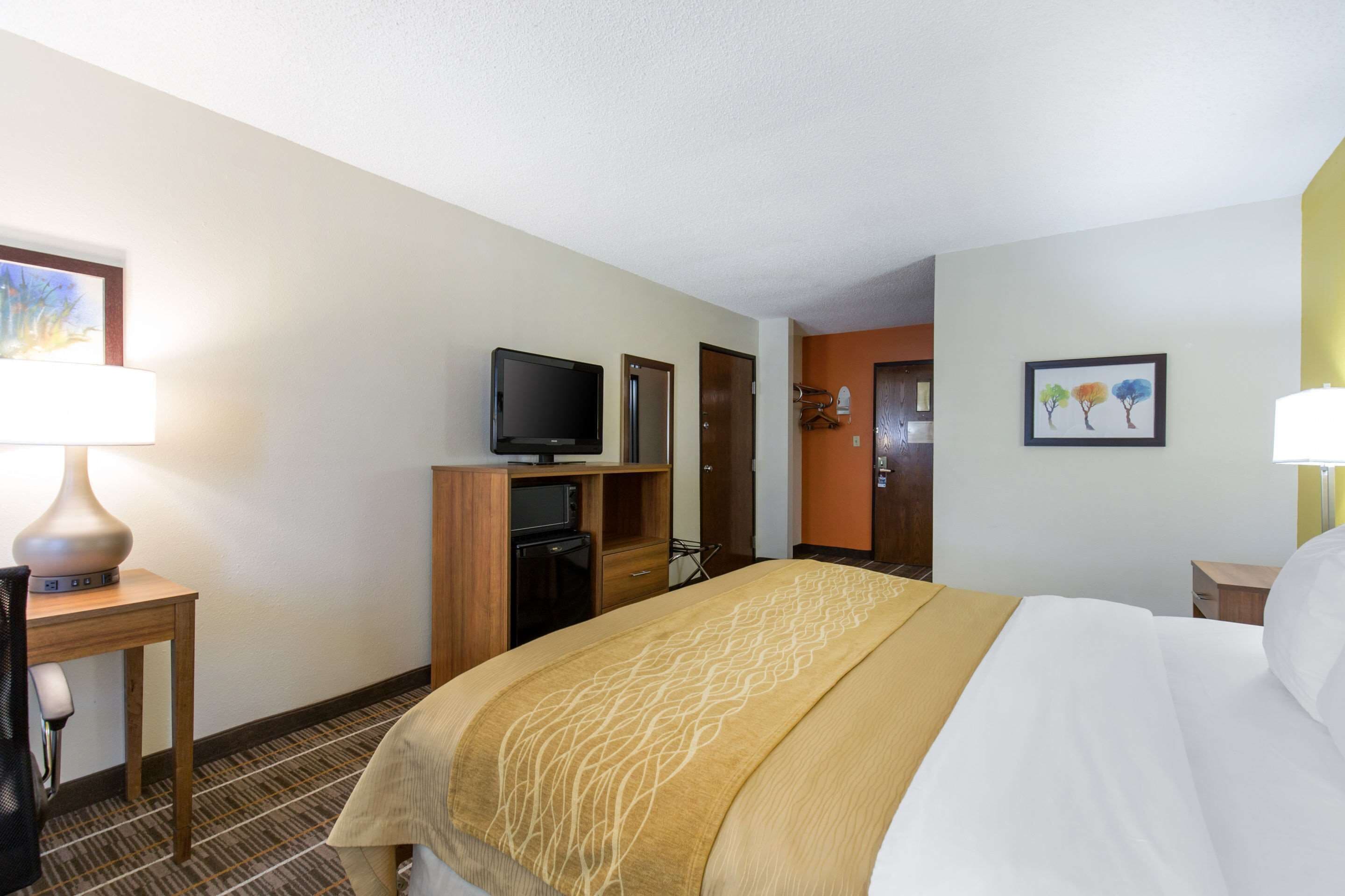 A hotel room with a large bed, a television on a wooden stand, a desk with a lamp, and a door leading to another room.
