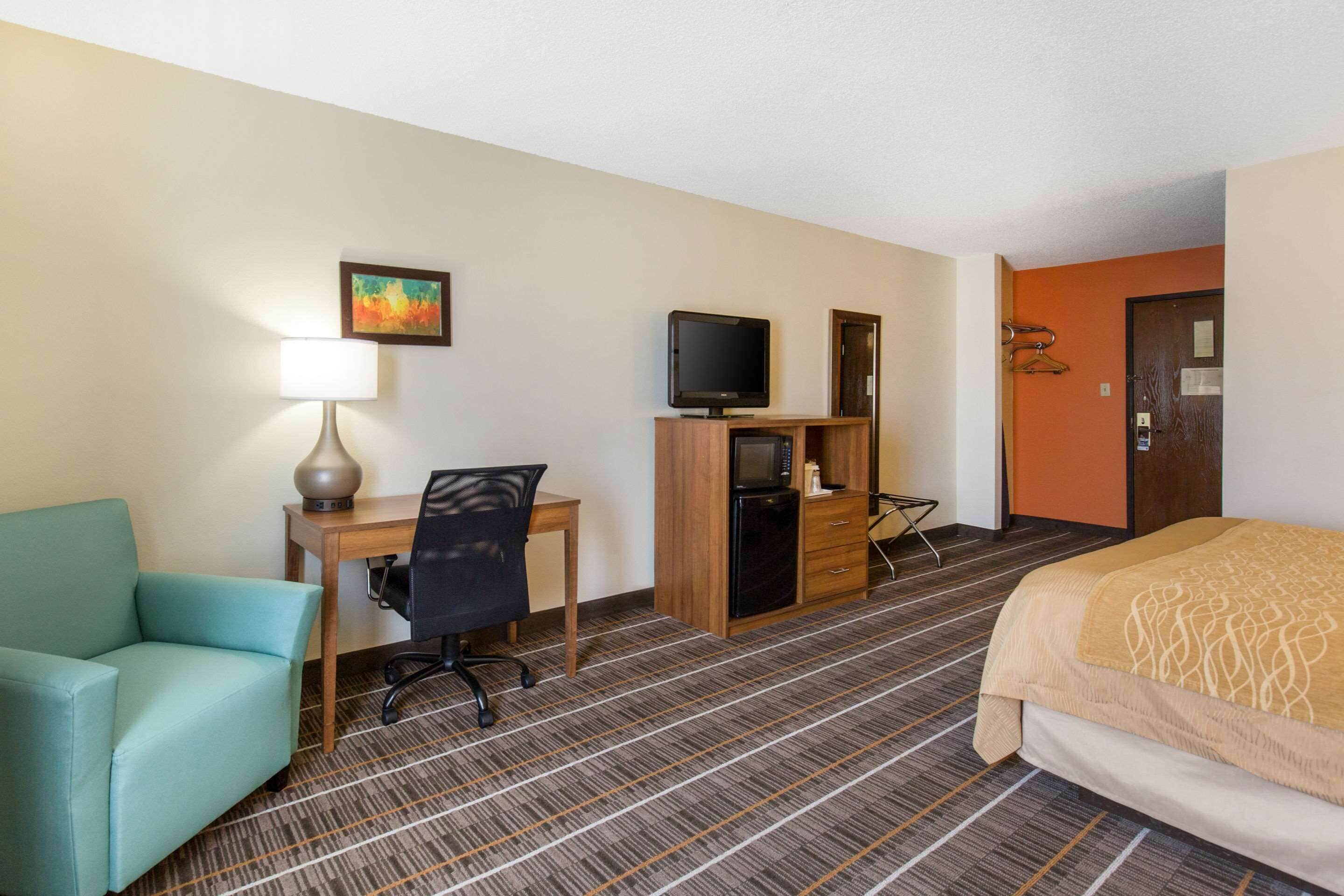 Una habitación de hotel con cama, escritorio con computadora, silla, lámpara, televisión y microondas.