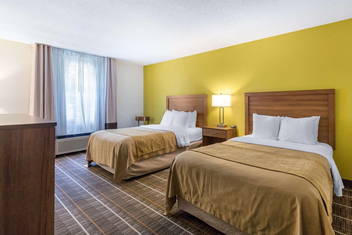 A hotel room with two beds, a desk, a lamp, and a window with curtains.