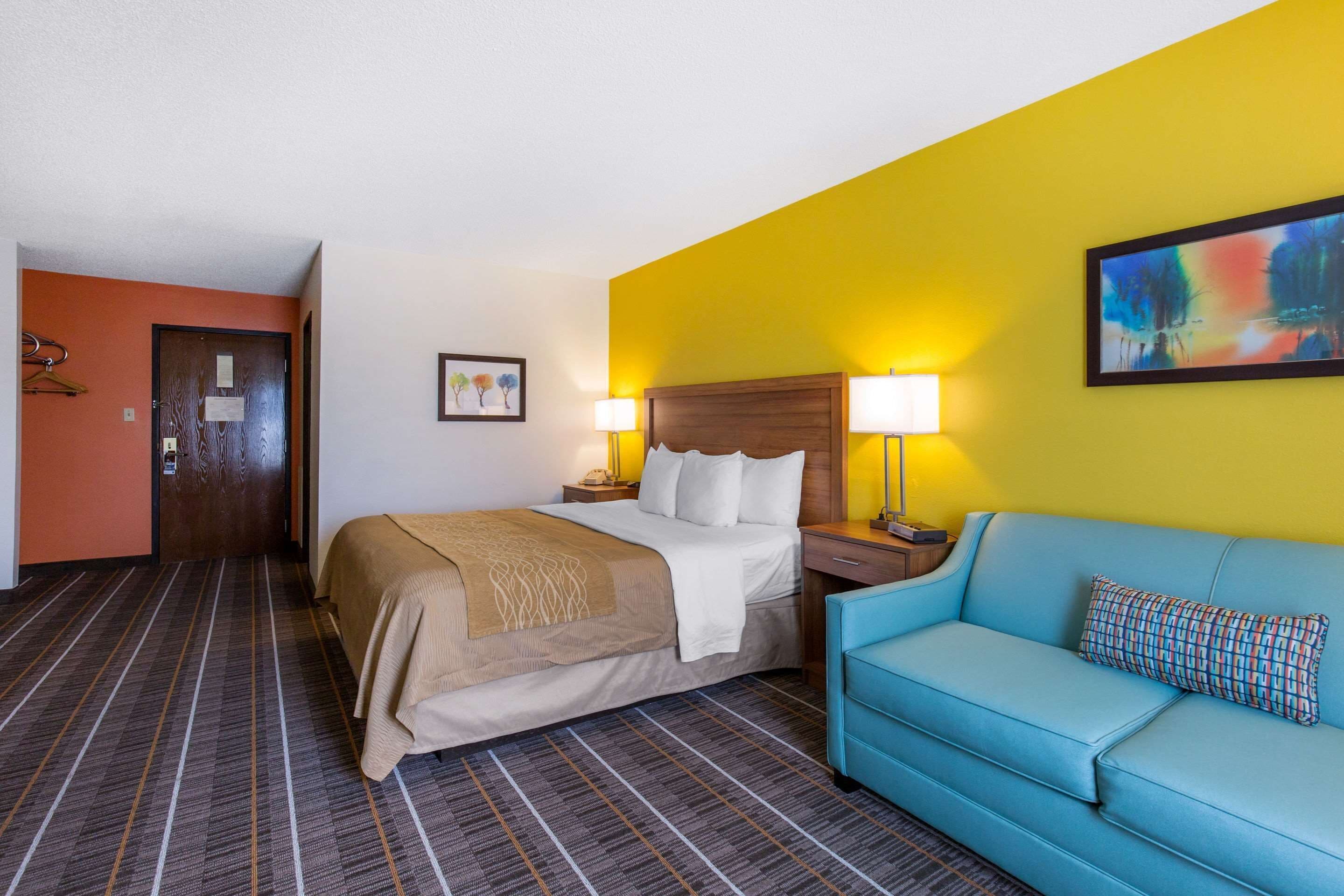 A hotel room with a bed, a blue sofa, a wooden headboard, a nightstand with lamps, and a painting on the wall.