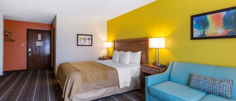 A hotel room with a bed, a blue sofa, a wooden headboard, a nightstand with lamps, and a painting on the wall.