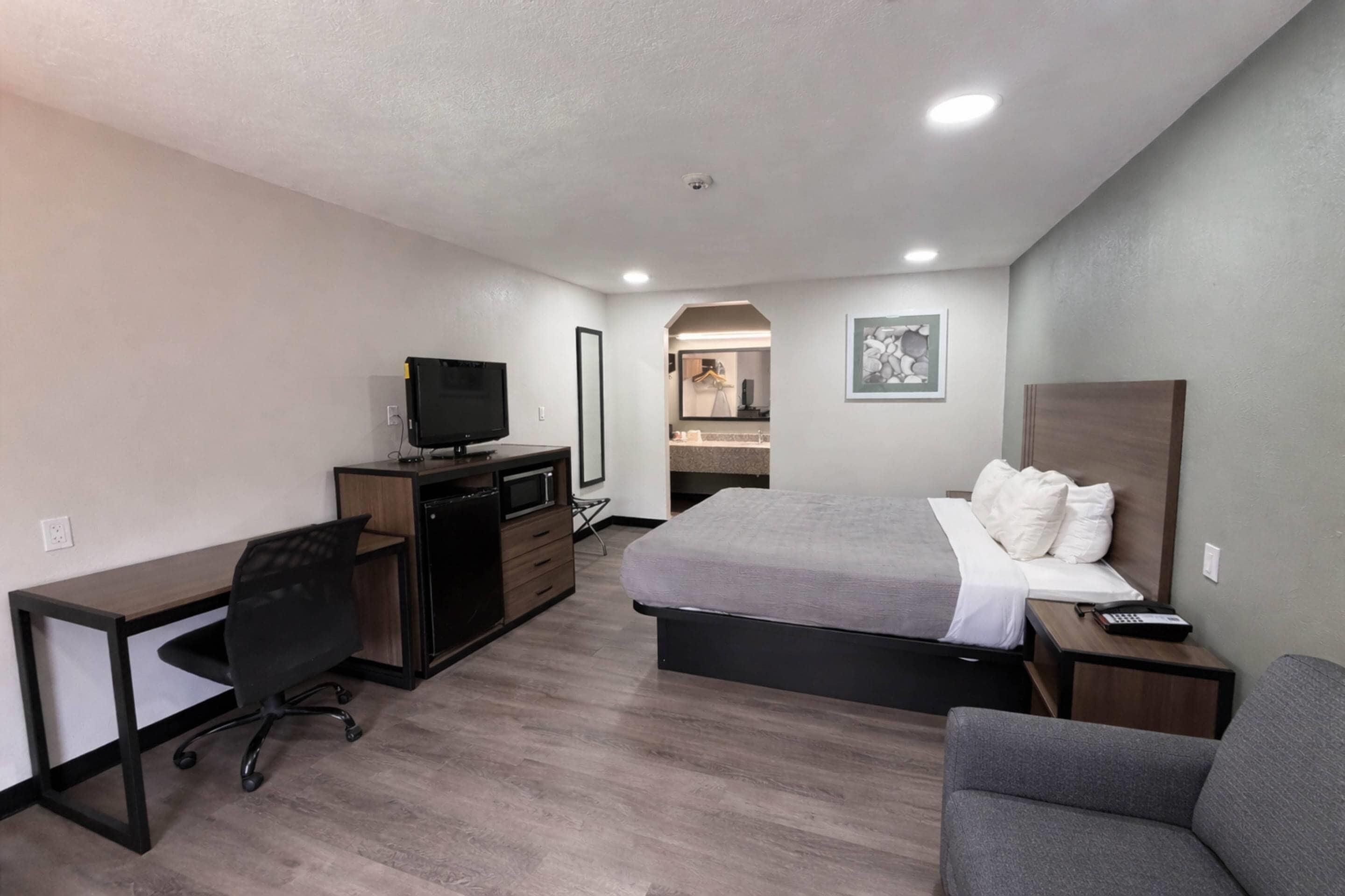 Habitación de hotel con cama, escritorio, silla, televisor, cómoda y baño visible a través de un arco.