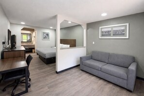 A modern hotel room with a desk, chair, grey sofa, two beds, and artwork on the walls.