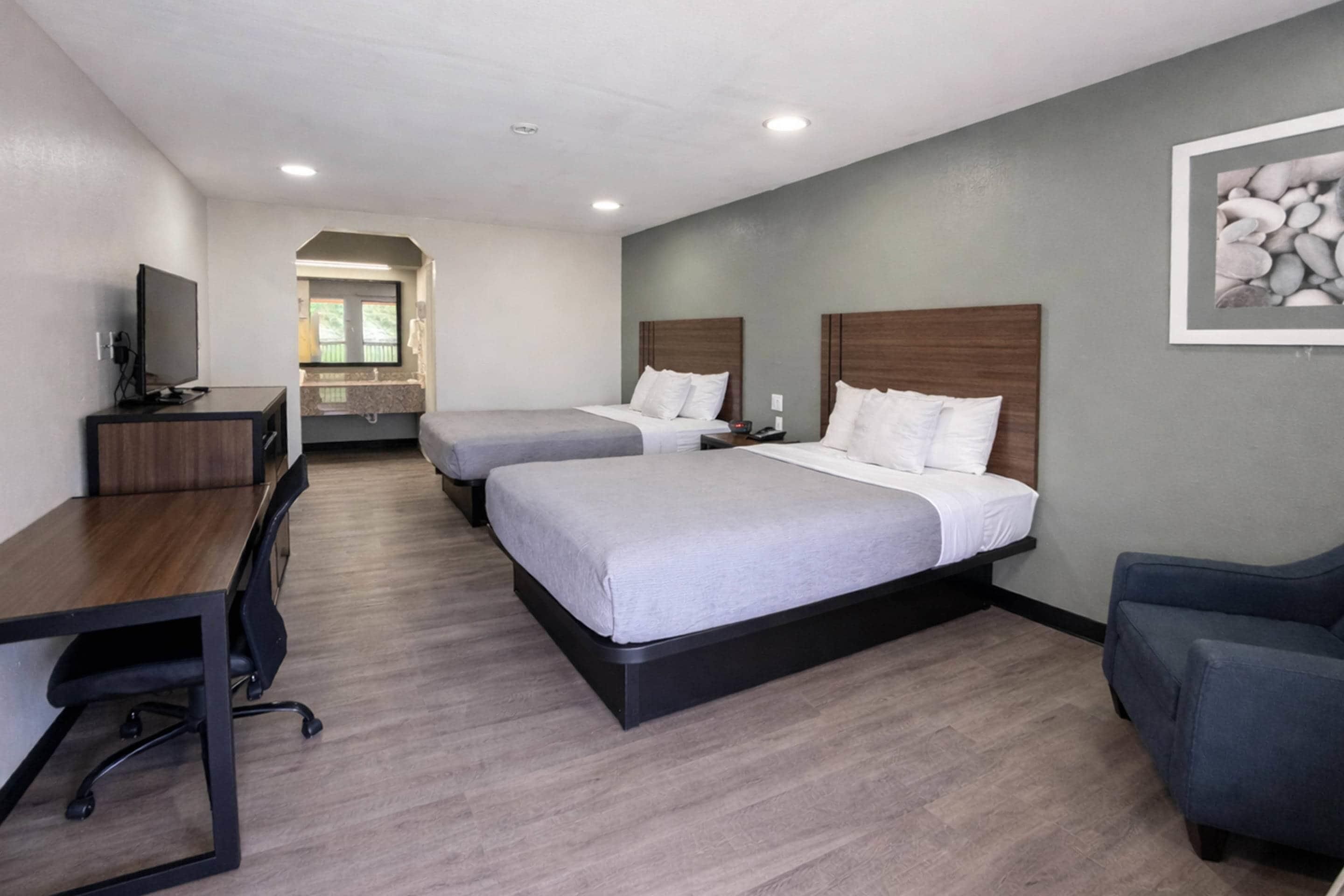 A hotel room with two beds, a desk, a chair, a television, and a bathroom visible through an archway.