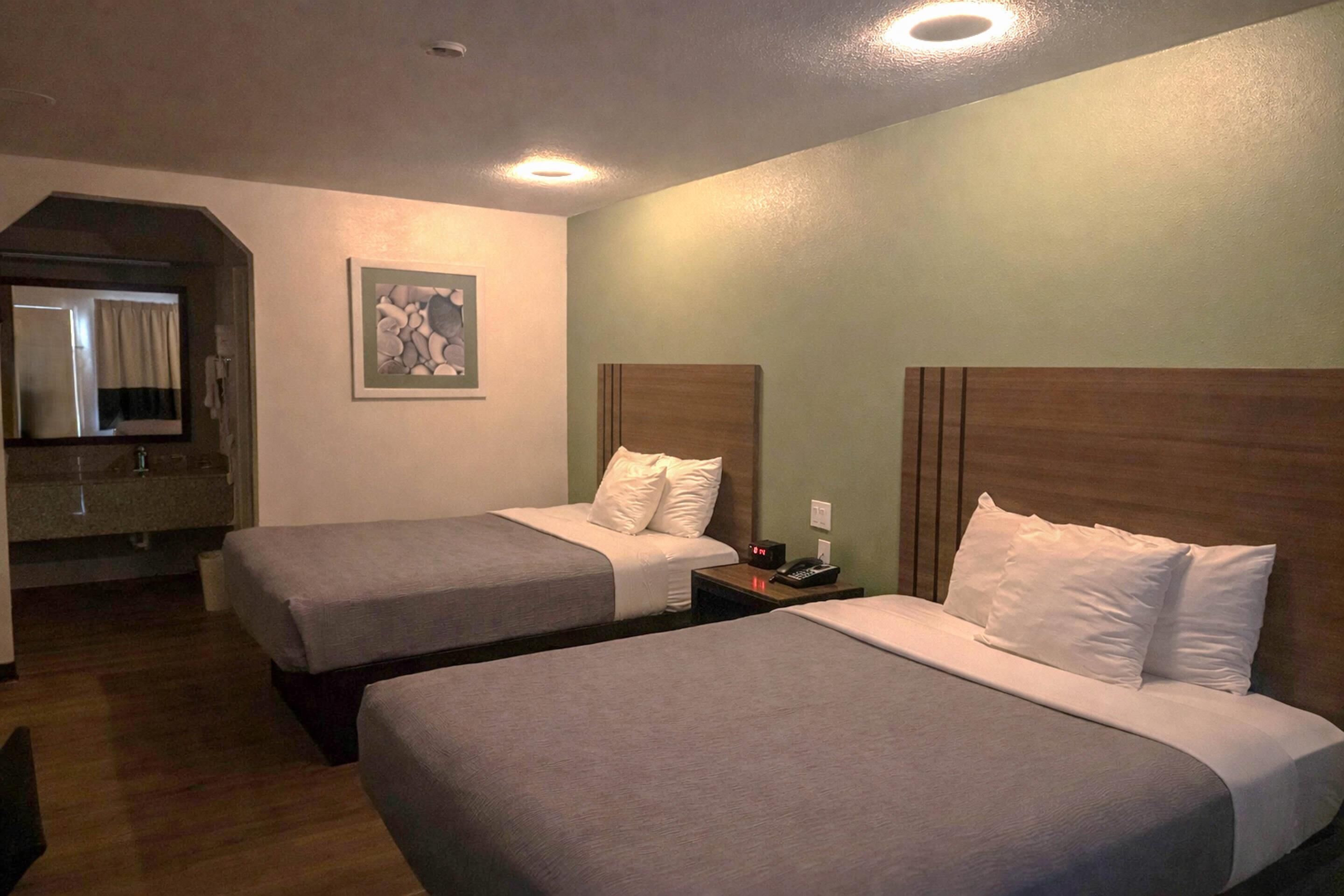 Habitación de hotel con dos camas, una mesita de noche con teléfono y un cuadro en la pared, además de un baño visible a través de un arco.