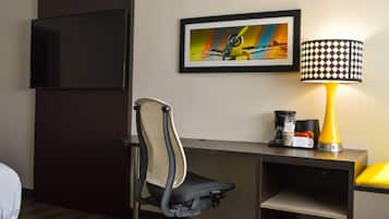 Quarto de hotel com uma escrivaninha, uma cadeira, televisão, quadro emoldurado, luminária e cafeteira.