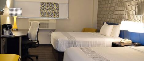 Habitación de hotel con dos camas, un escritorio con lámpara, una silla, lámpara de escritorio, teléfono y pared con diseño geométrico.