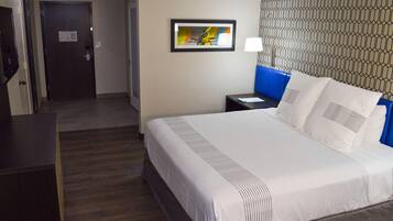 A hotel room with a large bed, a desk, a chair, a nightstand, a lamp, and a framed picture on the wall.