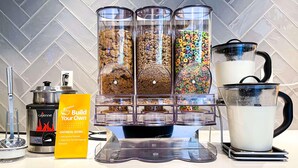 Un angolo colazione a buffet con distributori di porridge, cereali e latte, una macchina per il caffè e un cartello che indica 'Prepara la tua ciotola di porridge'.