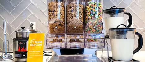Una estación de desayuno autoservicio con avena, cereales y dispensadores de leche, una máquina de café y un letrero que dice 'Prepara tu propia taza de avena'.