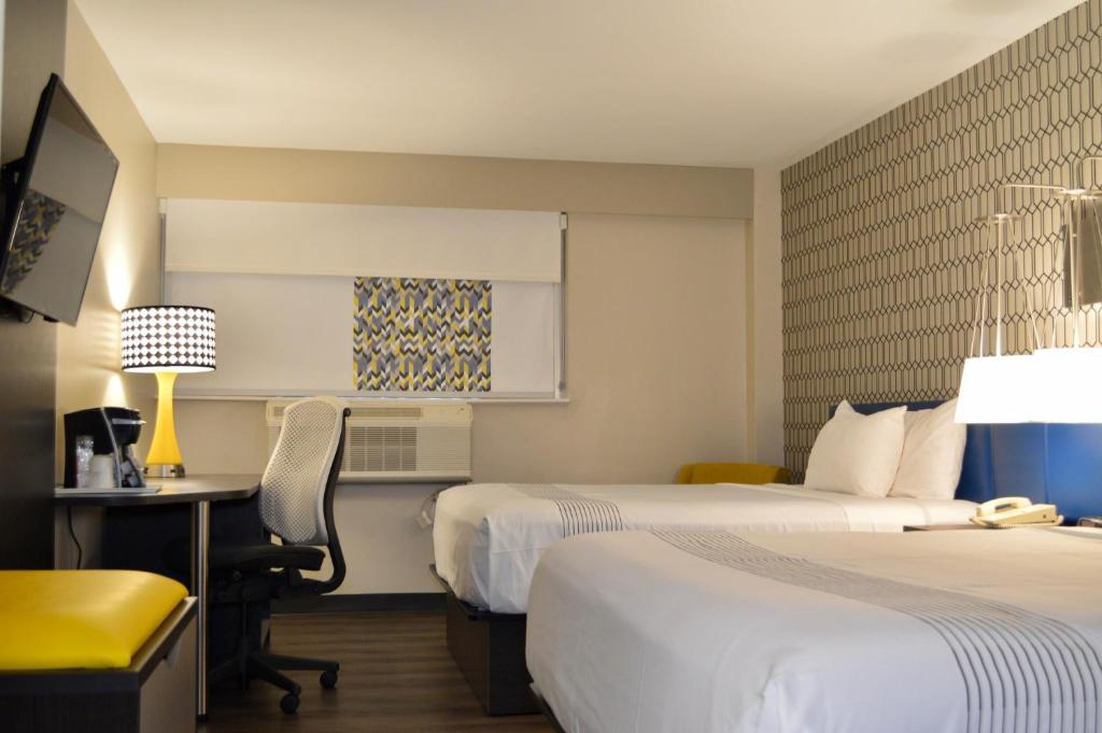 A hotel room with two beds, a desk with a lamp, a chair, a TV mounted on the wall, and a patterned artwork above the desk.