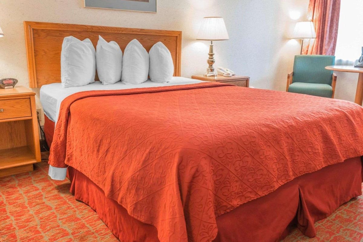 A hotel room with a large bed, a wooden headboard, a nightstand with a lamp, a chair, and a painting on the wall.