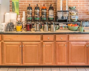 Una barra de desayuno con máquinas de café, un frutero y un dispensador de leche.