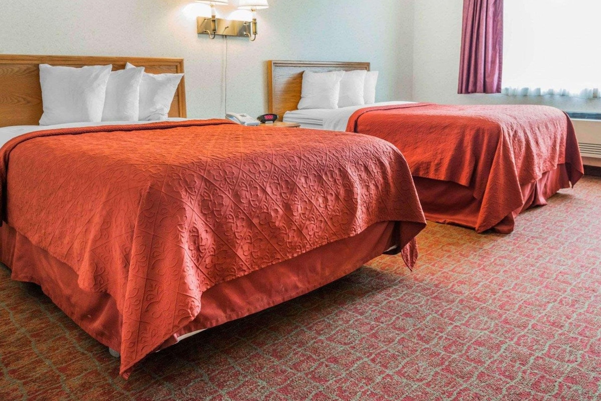 A hotel room with two beds, each with a red bedspread, a nightstand with a phone, and a wall-mounted lamp.