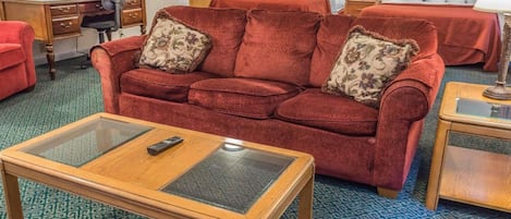Habitación de hotel con un sofá rojo, una mesa de centro de madera, un escritorio con una silla y dos camas con ropa de cama roja.