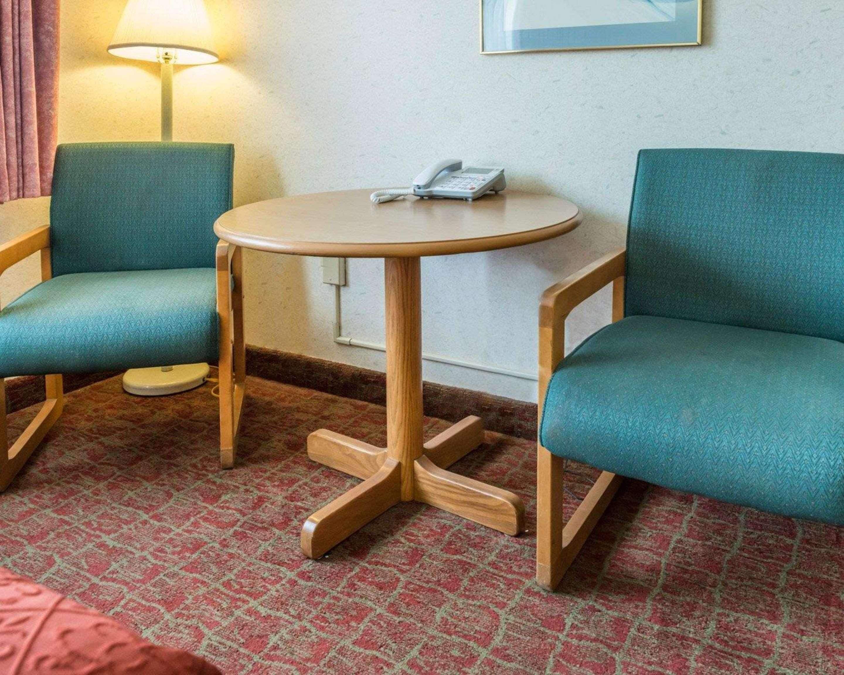 A hotel room with two teal upholstered chairs, a small round table with a telephone, a lamp, and a framed picture on the wall.
