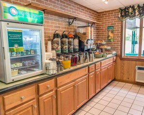 A breakfast bar with a refrigerator displaying items available during breakfast hours, a coffee station with multiple coffee makers, and a window with a view of the outdoors. - Quality Inn Royle (Kittanning)