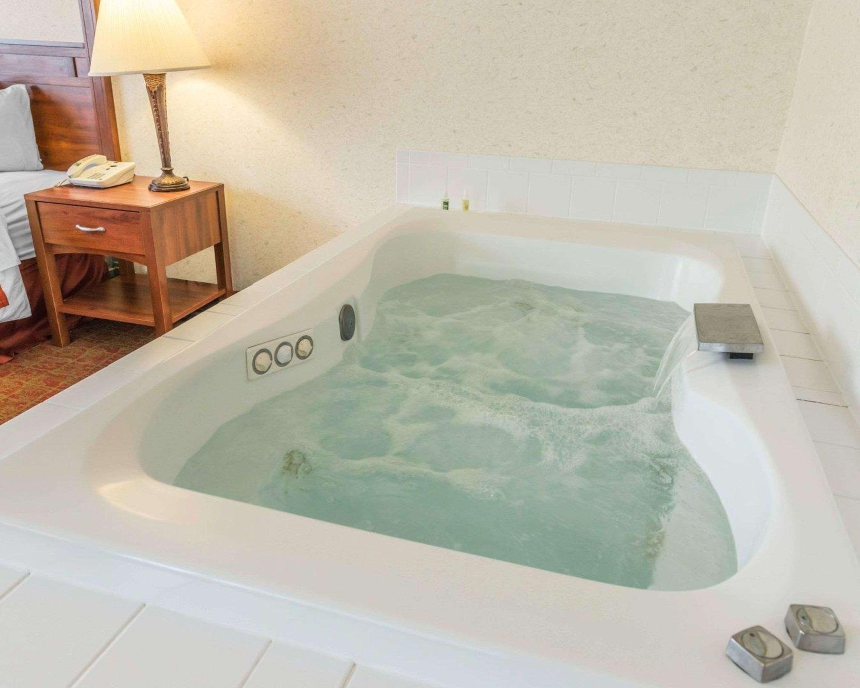 A hotel room with a large white bathtub filled with water, a wooden bedside table with a lamp, a phone, and a small drawer.