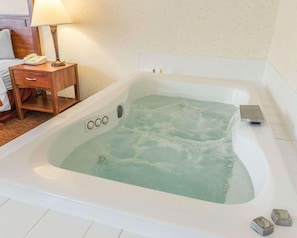 Quarto de hotel com uma banheira grande e branca cheia de água, mesa de cabeceira de madeira com abajur, telefone e pequena gaveta.