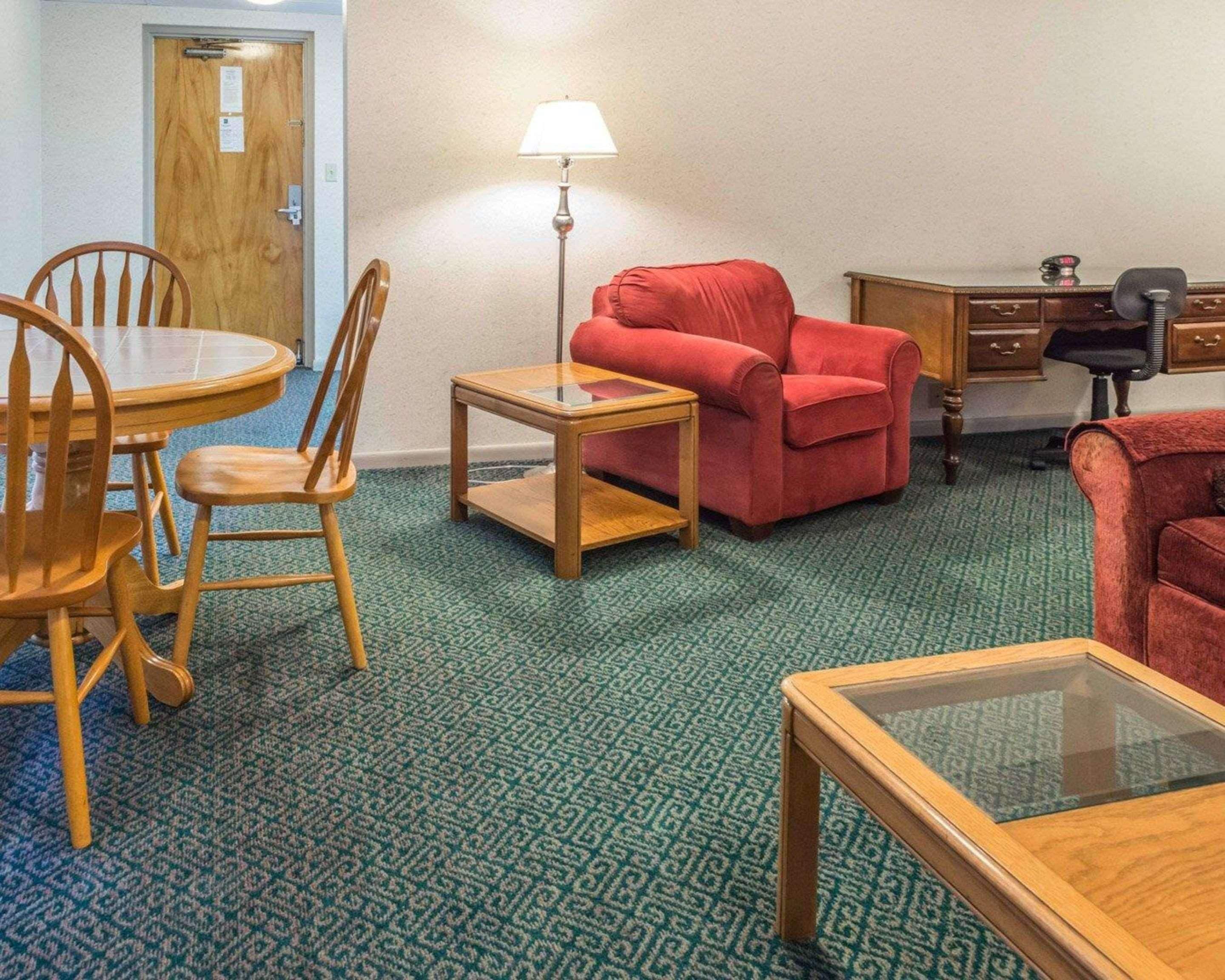 A room with a wooden dining table, two chairs, a red sofa, a small wooden coffee table, a lamp, a desk with a chair, and a patterned carpet.