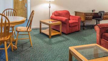 A room with a wooden dining table, two chairs, a red sofa, a small wooden coffee table, a lamp, a desk with a chair, and a patterned carpet.