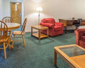 A room with a wooden dining table, two chairs, a red sofa, a small wooden coffee table, a lamp, a desk with a chair, and a patterned carpet. - Quality Inn Royle (Kittanning)