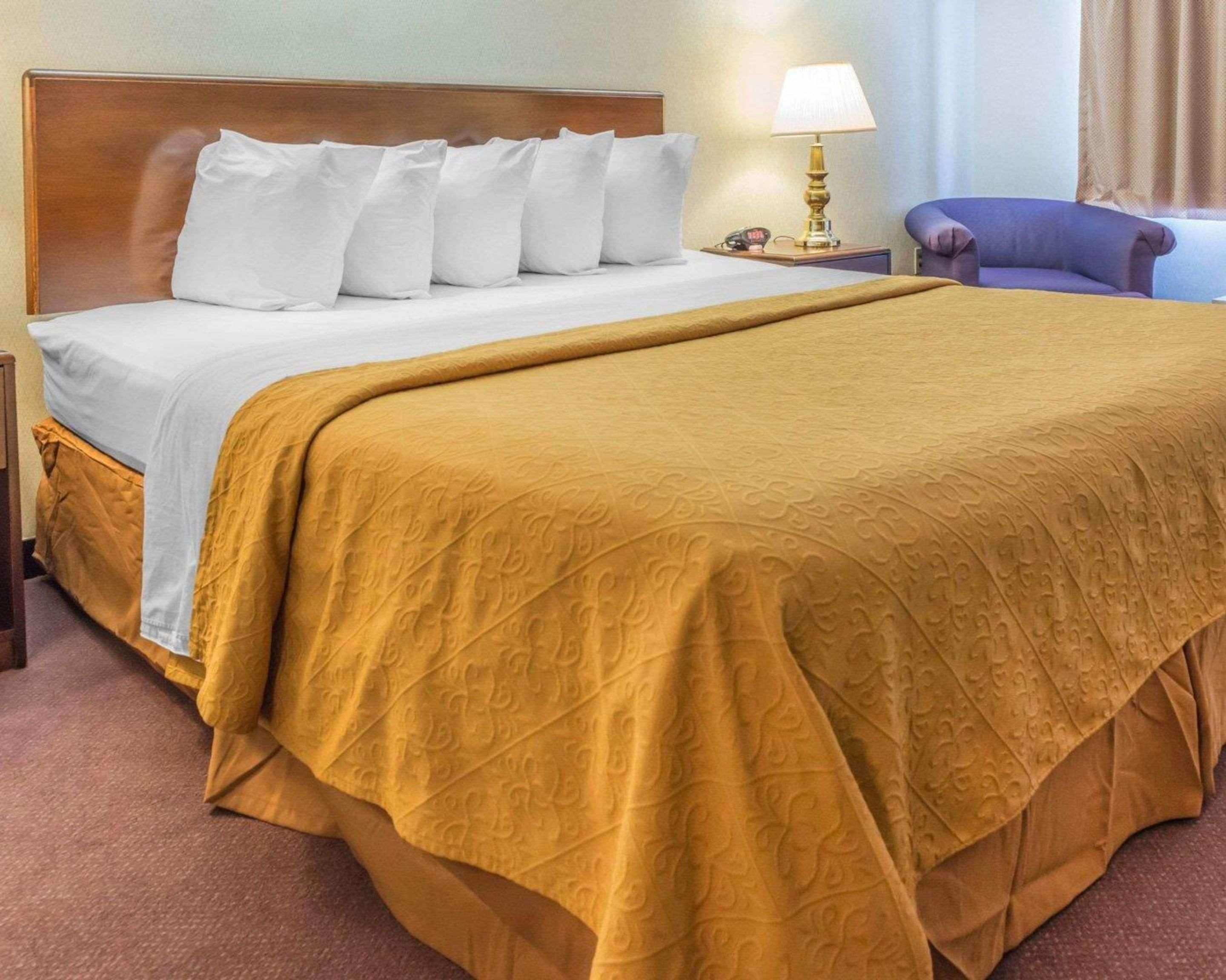 Quarto de hotel com uma cama grande, cabeceira de madeira, travesseiros brancos, edredom bege-mostarda, luminária de cabeceira e poltrona roxa.