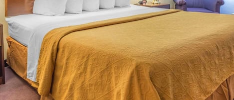 A hotel room with a large bed, a wooden headboard, white pillows, a mustard-colored bedspread, a bedside lamp, and a purple armchair.