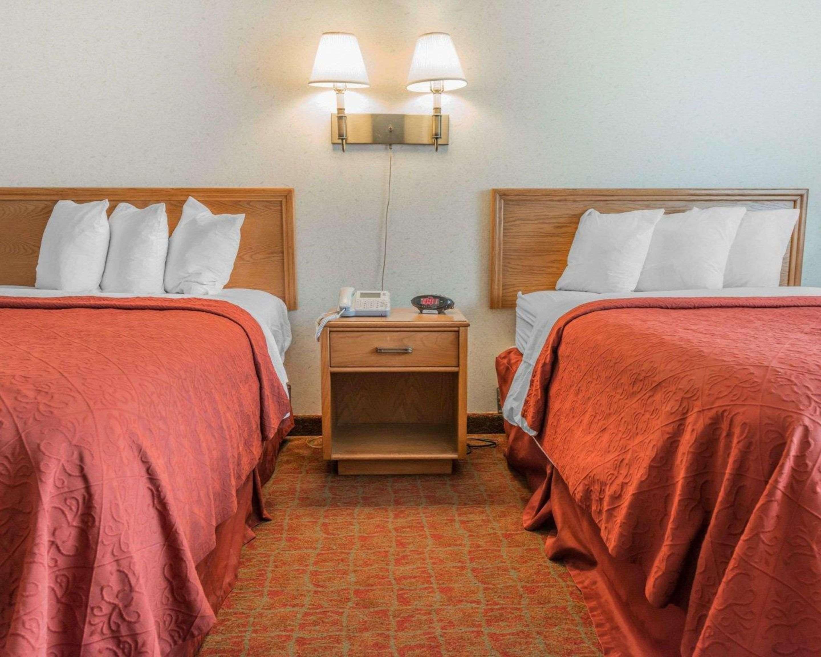 A hotel room with two beds, each with a wooden headboard, a nightstand with a phone and a clock, and a wall-mounted lamp.