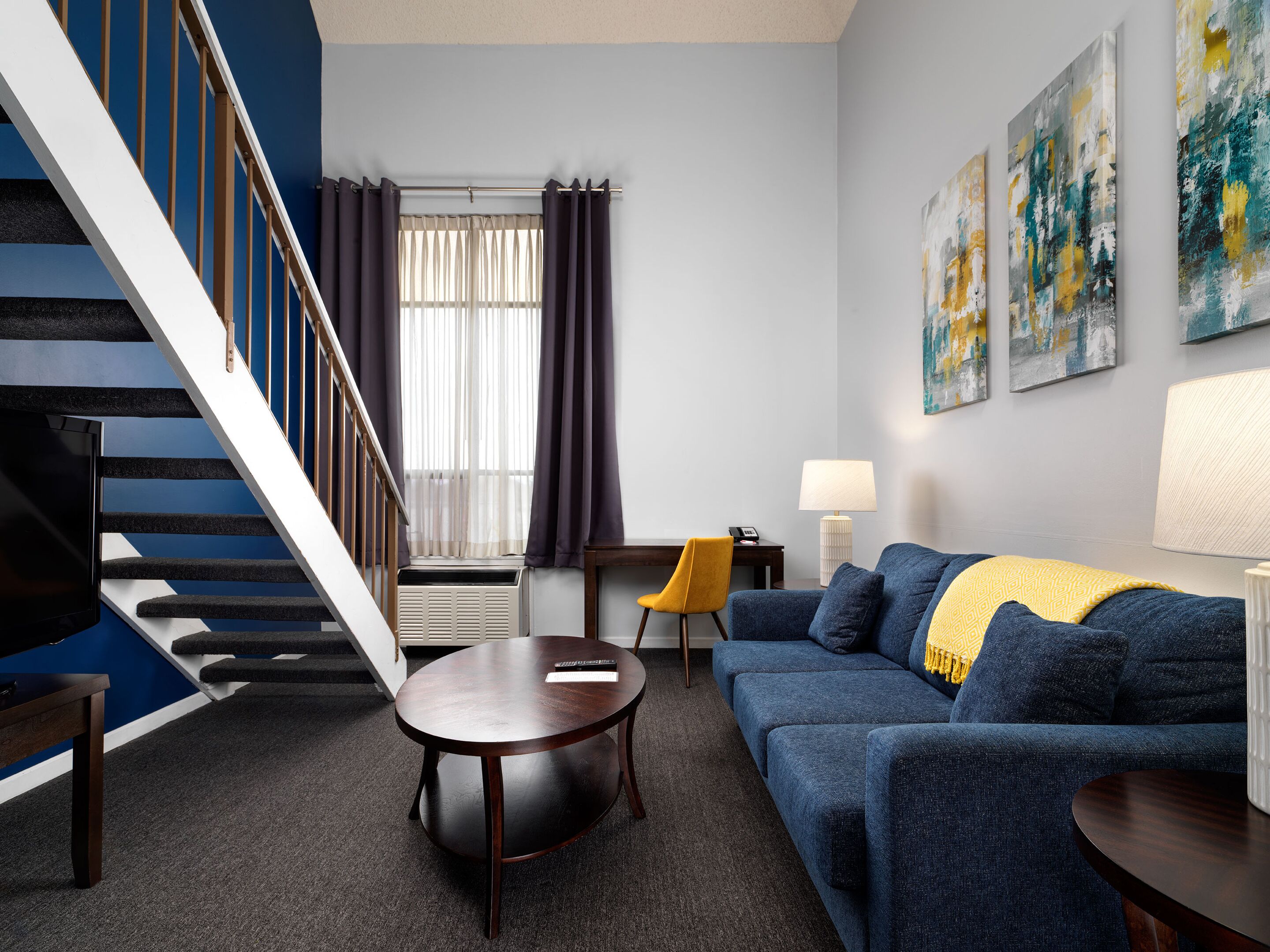 A modern living room with a blue sofa, a round wooden coffee table, a yellow chair, a lamp, and a staircase with a railing.