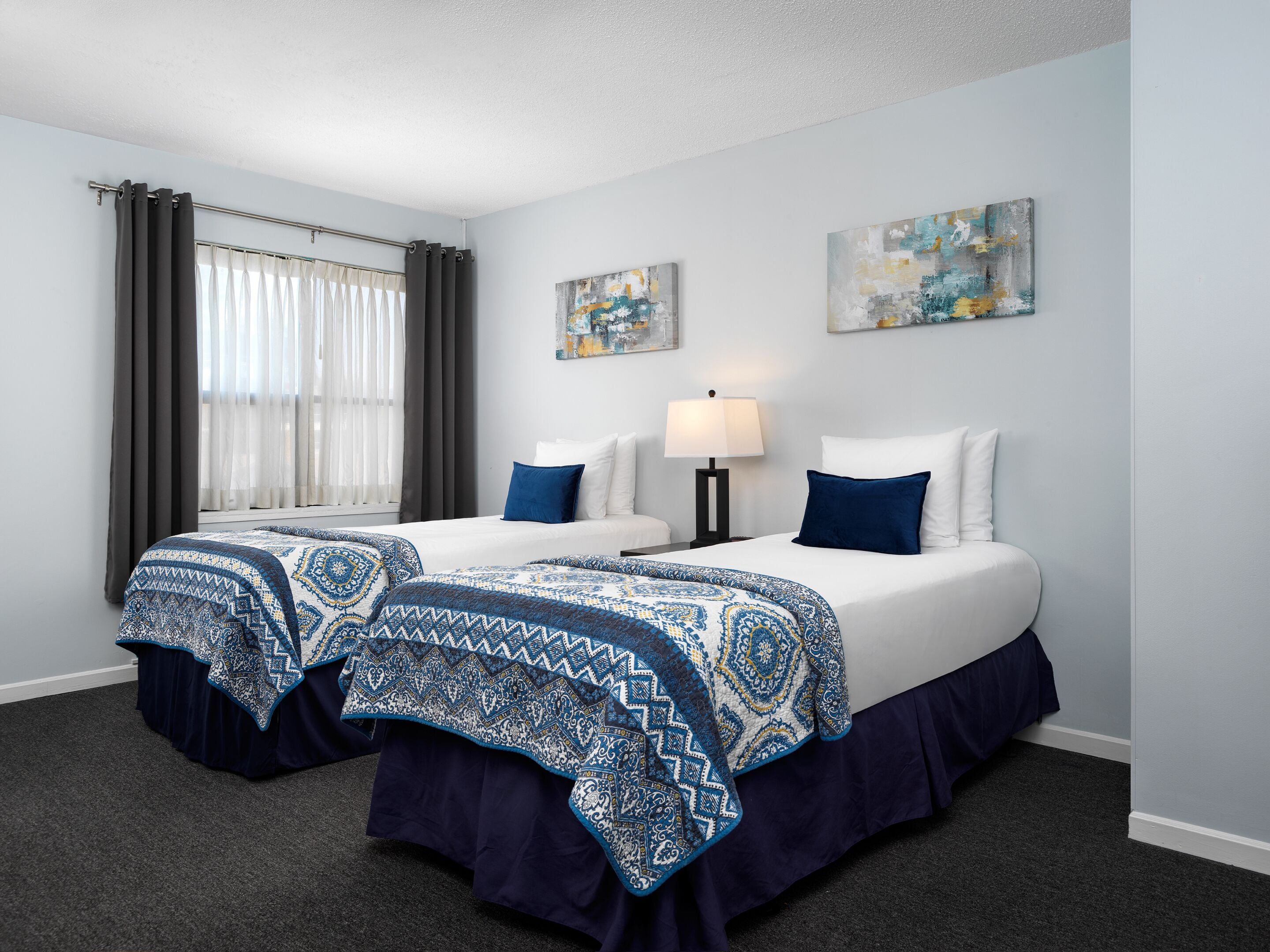A hotel room with two beds, dark blue and white bedding, a window with sheer curtains, and two abstract paintings on the wall.