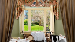 Comedor con mesas dispuestas para cuatro, un ventanal con vistas a un jardín y una araña colgante.
