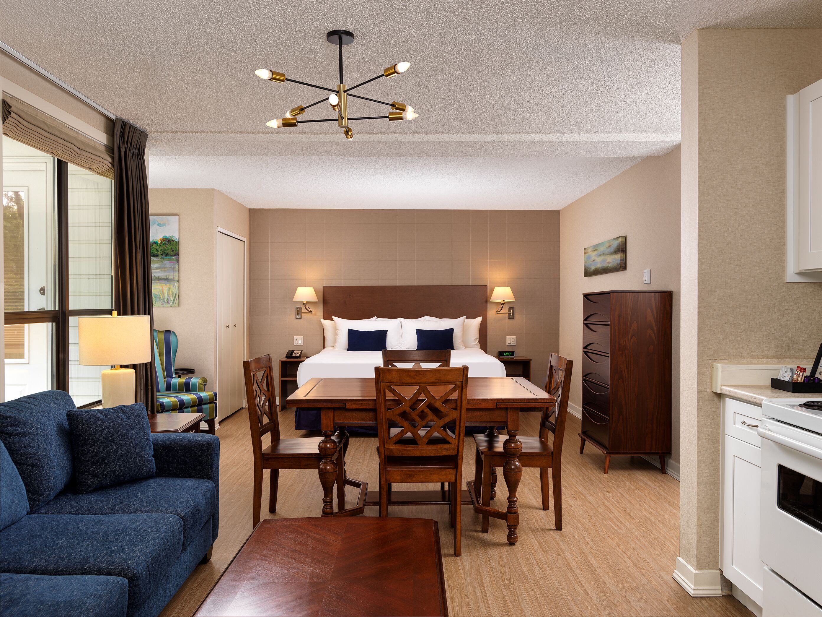 A hotel room with a bed, bedside lamps, a dining table with chairs, a sofa, a wooden cabinet, and a kitchen area.