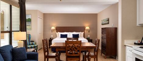 A hotel room with a bed, bedside lamps, a dining table with chairs, a sofa, a wooden cabinet, and a kitchen area.