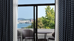 Un balcón con sillas de mimbre y una mesa, con vista a un río que incluye un puente y vegetación.