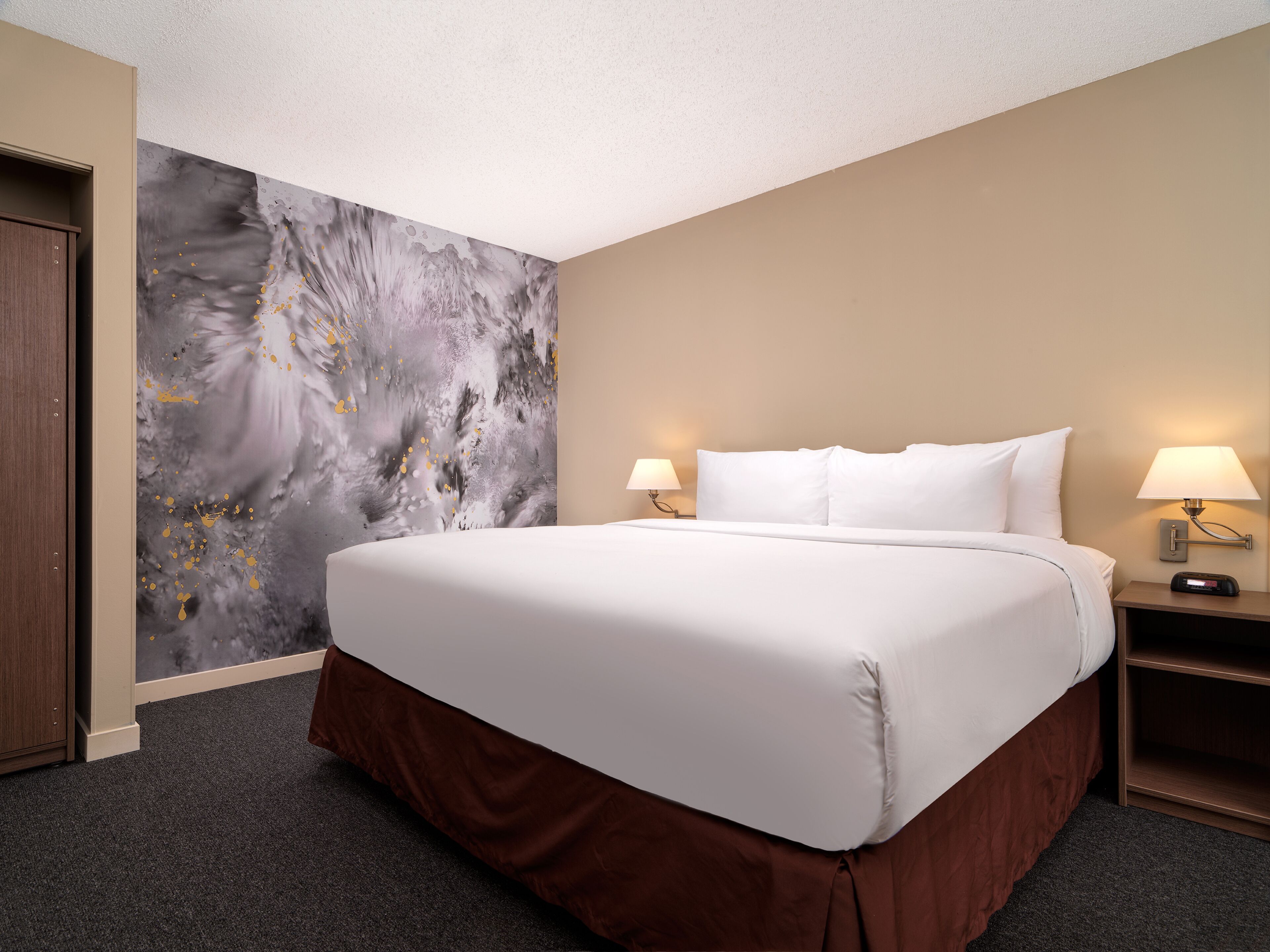 Una habitación de hotel con una cama grande, dos mesitas de noche con lámparas, una pared con un cuadro abstracto de gran tamaño y una puerta.