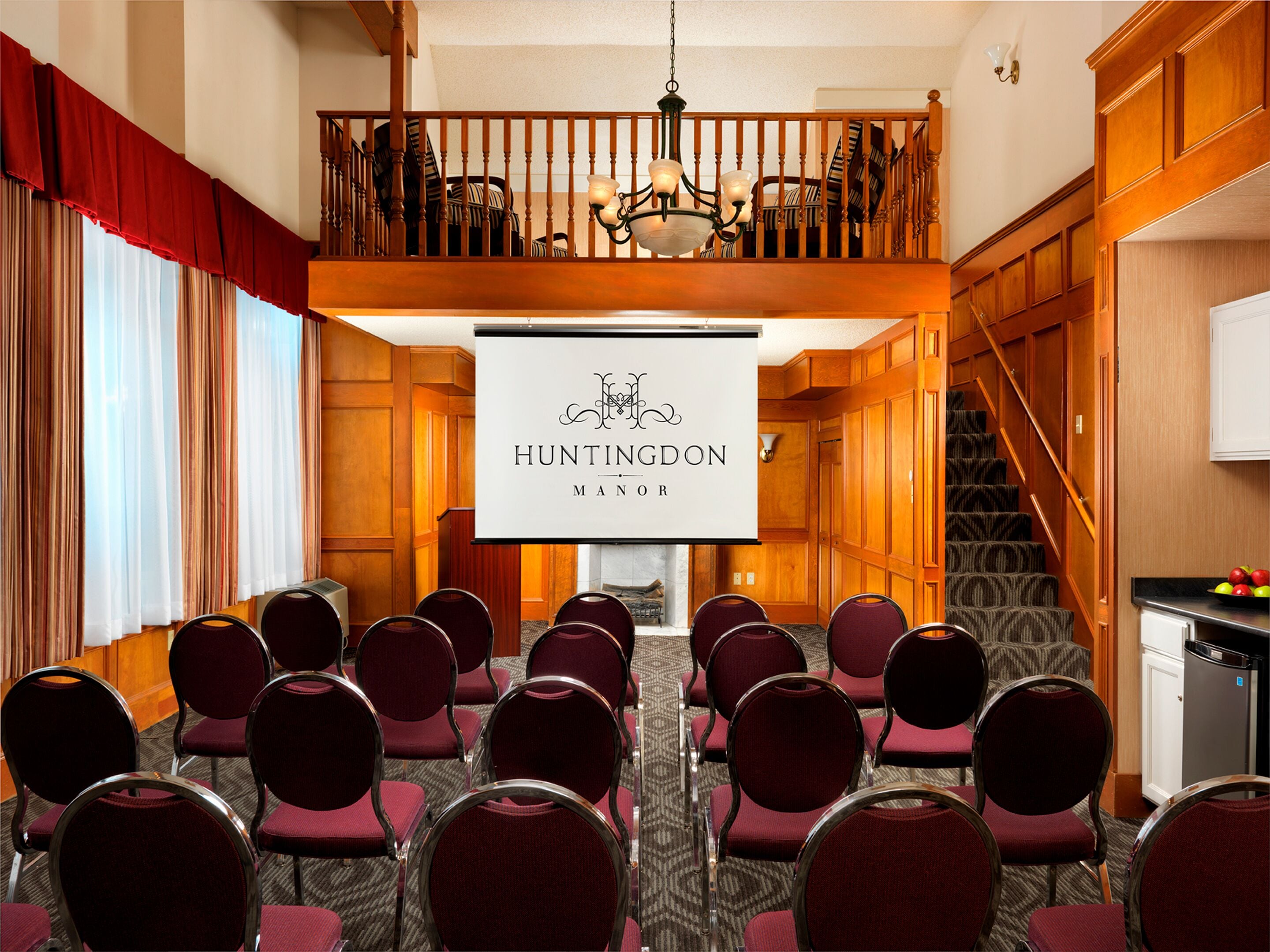 Un salón de conferencias con filas de sillas, una pantalla de proyector mostrando "Huntingdon Manor", una araña, paneles de madera y una escalera.