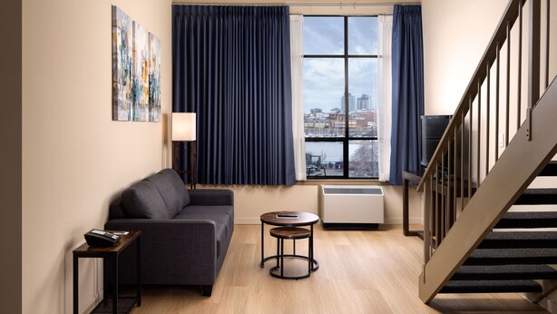 A modern living room with a sofa, a small table, a lamp, and a view of the city through large windows.