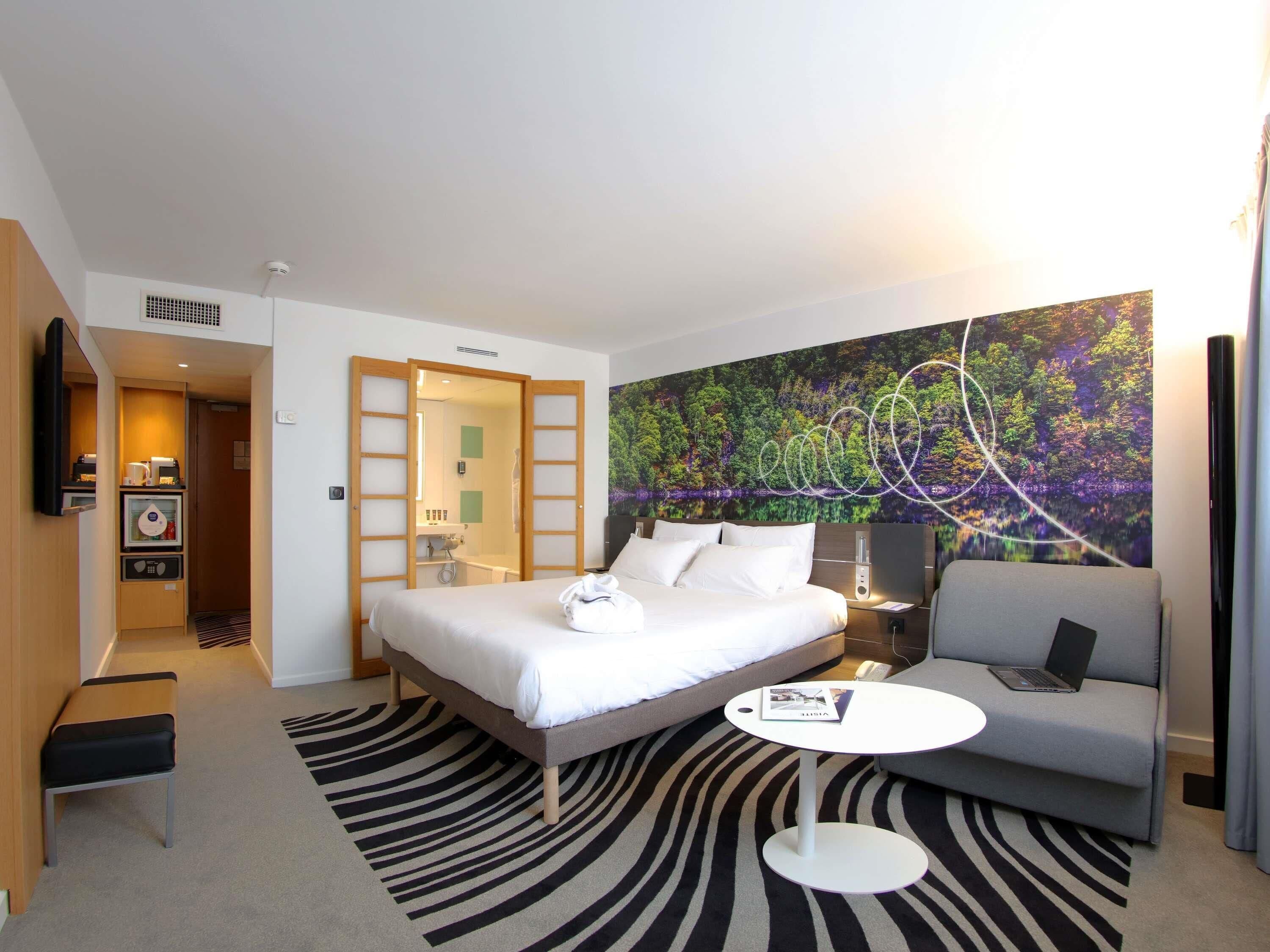 A hotel room with a bed, a sofa, a small table, a bathroom with a shower, and a large forest-themed wall mural.