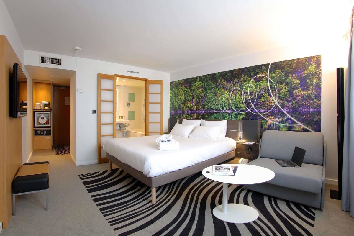 A hotel room with a bed, a sofa, a small table, a bathroom with a shower, and a large forest-themed wall mural.